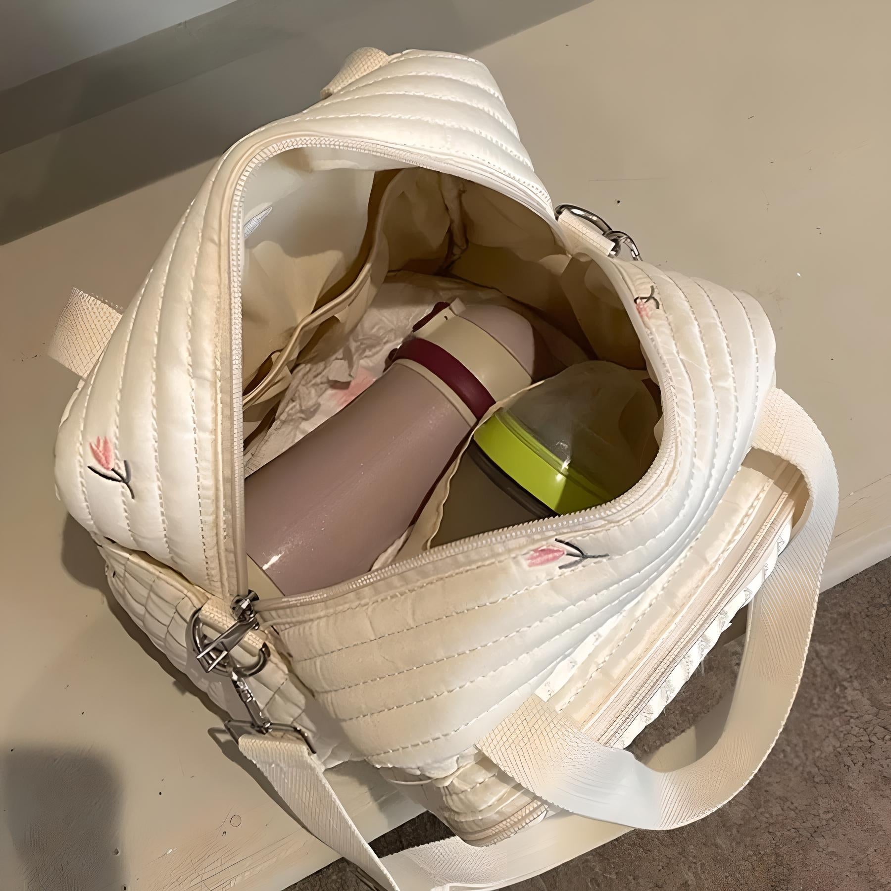 Quilted white tote bag with floral embroidery, open to reveal a pink thermos and a green-lidded container inside, on a beige surface.