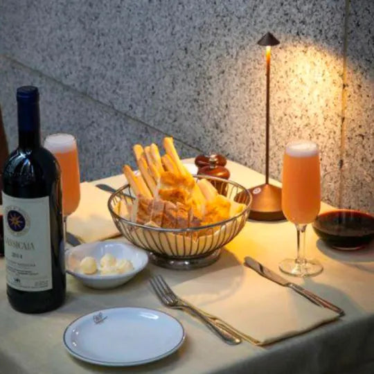 Elegant dining setup with a bottle of red wine, breadsticks in a basket, two glasses of orange juice, and a lit table lamp on a white tablecloth.