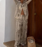 Woman wearing cactus print pajama set, standing by a wooden door, with a woven basket on the floor. Fashionable loungewear, cozy home style.