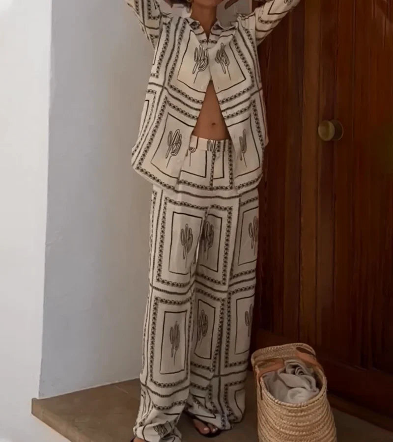 Woman wearing cactus print pajama set, standing by a wooden door, with a woven basket on the floor. Fashionable loungewear, cozy home style.