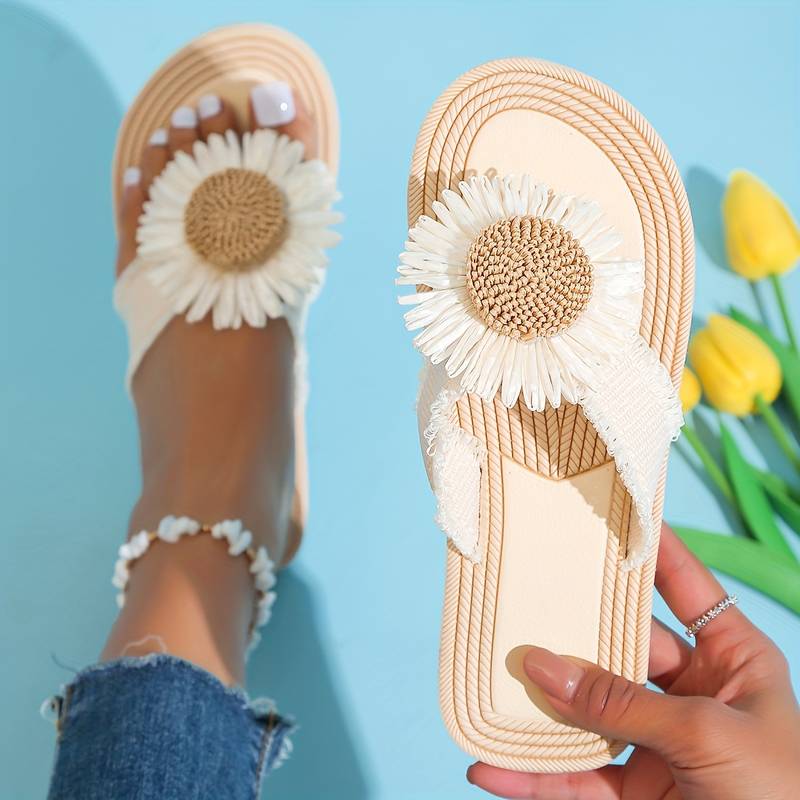 Beige sandals with large white daisy flower design, worn on a foot with painted toenails, against a blue background with yellow tulips. Summer footwear.