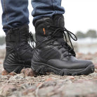 Black tactical combat boots on rocky terrain, worn with blue jeans. Durable, high-ankle design ideal for outdoor activities and rugged environments.