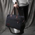 Stylish black canvas duffel bag with brown leather straps, held by a person in casual attire. Perfect for travel, gym, or everyday use.