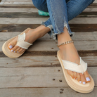 Woman wearing frayed hem jeans and beige platform flip-flops with white toenail polish, sitting on wooden deck. Fashionable summer footwear.