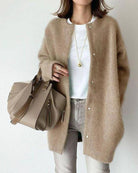 Woman in beige cardigan and white shirt holding a taupe leather handbag. Fashionable casual outfit with gold necklace and light jeans.