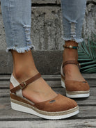 Women's brown suede espadrille wedges with ankle strap, paired with distressed denim jeans. Stylish summer footwear on wooden floor background.