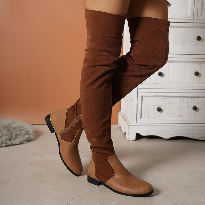 Brown over-the-knee suede boots with side zipper, flat sole, and leather accents, styled indoors on a beige floor near a white wooden dresser.