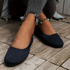 Women's navy blue knit ballet flats on wooden floor, paired with frayed hem jeans and a turquoise anklet. Comfortable, stylish footwear for casual wear.