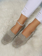 Women's beige knit loafers on feet with white distressed jeans, pearl anklet, and fluffy rug background. Stylish casual footwear for women.