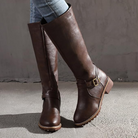 Brown knee-high leather boots with buckle detail, worn with jeans, on a concrete floor. Stylish women's footwear, perfect for fall fashion.