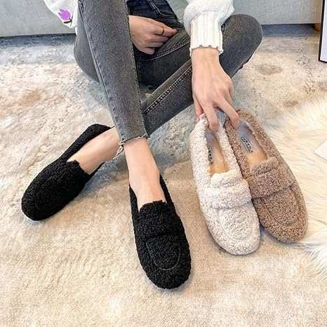 Woman holding cozy faux fur loafers in black and beige, sitting on a plush carpet. Casual winter footwear, stylish and comfortable slip-on shoes.