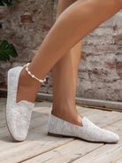 White woven slip-on loafers on woman's feet, featuring a pearl anklet, set against a rustic brick wall and wooden floor. Casual footwear fashion.