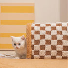 White kitten peeking from a checkered brown and white rug, with a yellow striped background. Cute pet, cozy home decor, playful cat.