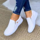 White slip-on sneakers with zipper detail on a woven rug, paired with distressed jeans and a gold anklet. Stylish casual footwear for women.