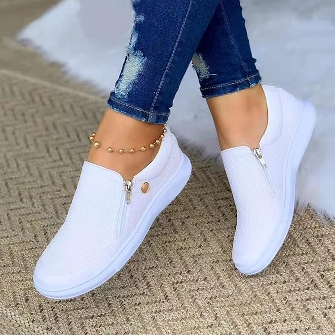 White slip-on sneakers with zipper detail on a woven rug, paired with distressed jeans and a gold anklet. Stylish casual footwear for women.