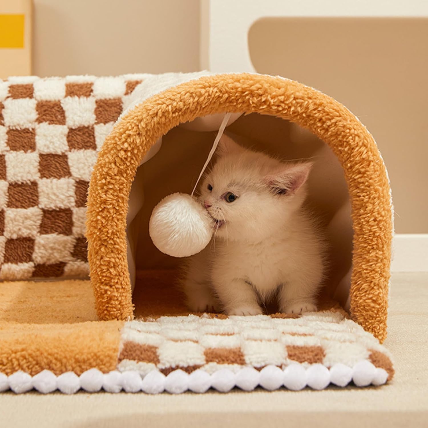 Fluffy white kitten playing with a hanging ball inside a cozy checkered cat tunnel. Perfect pet toy for indoor cats, enhancing playtime and comfort.