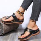 Women's black leather sandals with toe loop, worn with beaded anklets. Casual summer footwear on a wooden surface. Fashionable and comfortable design.