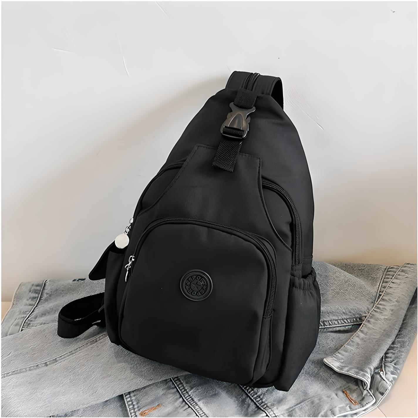 Black sling backpack with multiple compartments, adjustable strap, and zipper closures, placed on a denim jacket. Ideal for travel and daily use.