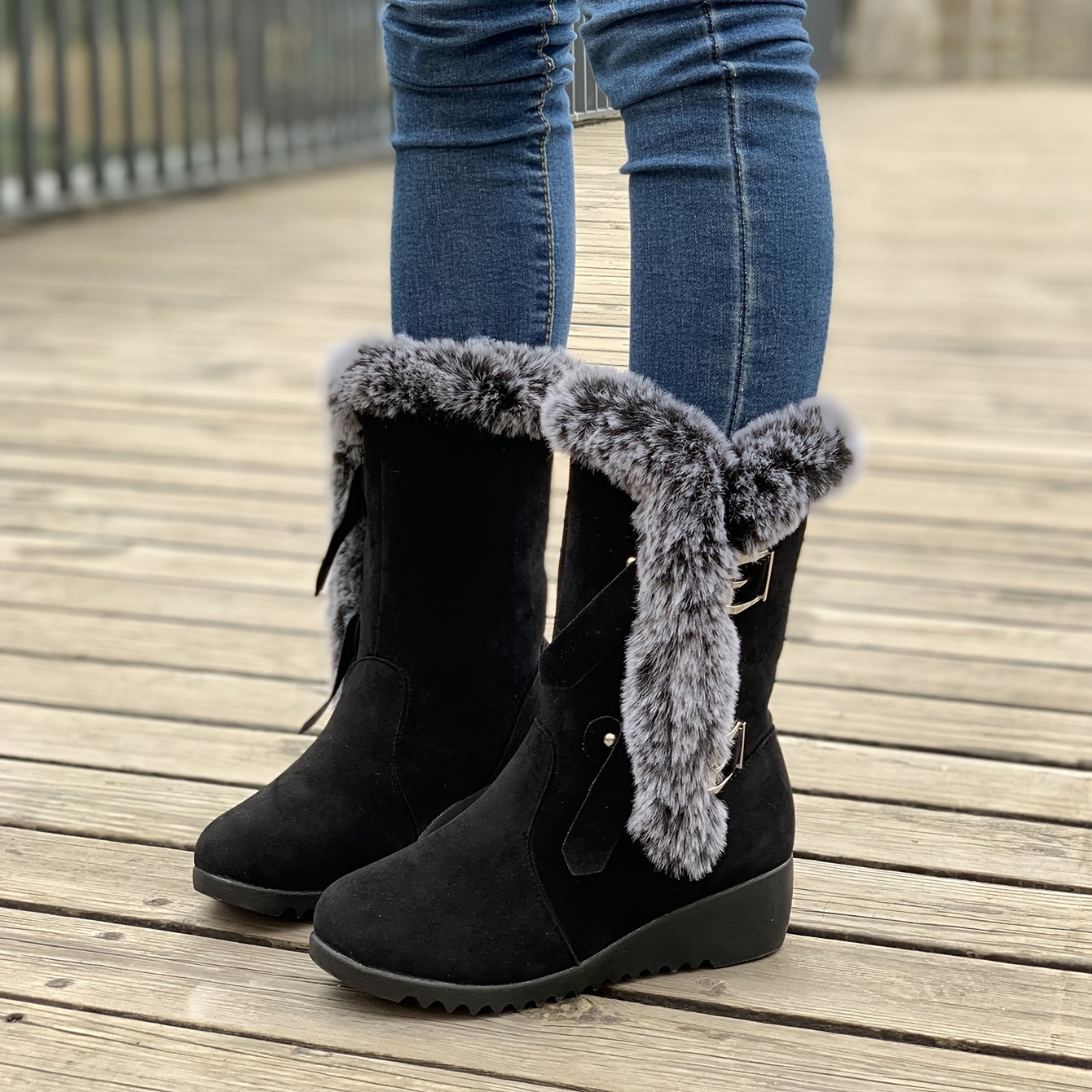 Black suede winter boots with faux fur trim and wedge heel, worn with blue jeans on a wooden deck. Fashionable women's footwear for cold weather.