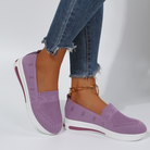 Purple slip-on sneakers with white soles, worn with frayed hem jeans and a gold anklet. Stylish women's casual footwear, comfortable knit design.