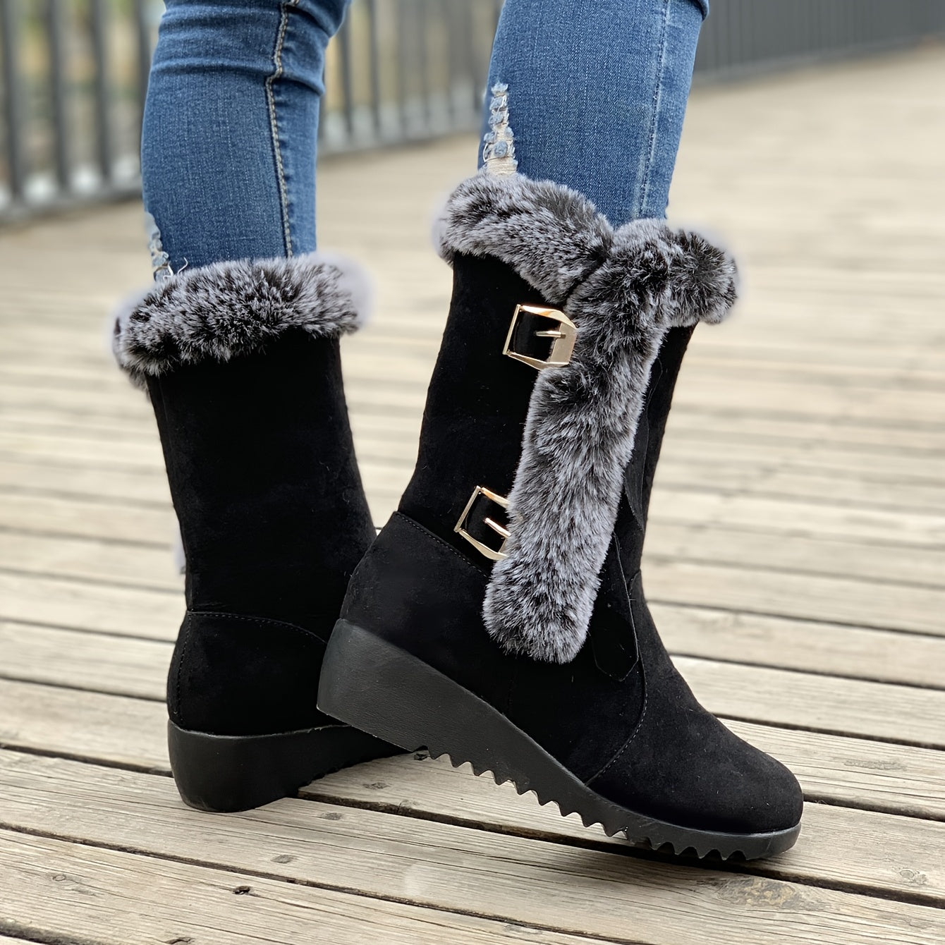 Black suede winter boots with faux fur trim and buckle detail, worn with blue jeans on a wooden deck. Stylish, warm footwear for cold weather.