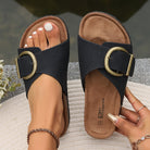 Black leather slide sandals with large gold buckles, worn by a person with manicured nails, on a textured surface. Fashion footwear, summer style.