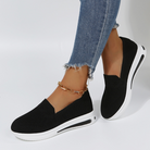 Black slip-on sneakers with white soles, worn with frayed hem jeans and a gold anklet. Stylish women's casual footwear, comfortable and trendy.