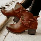 Brown leather ankle boots with block heels and decorative straps, worn with black jeans. Stylish women's footwear for casual and trendy outfits.