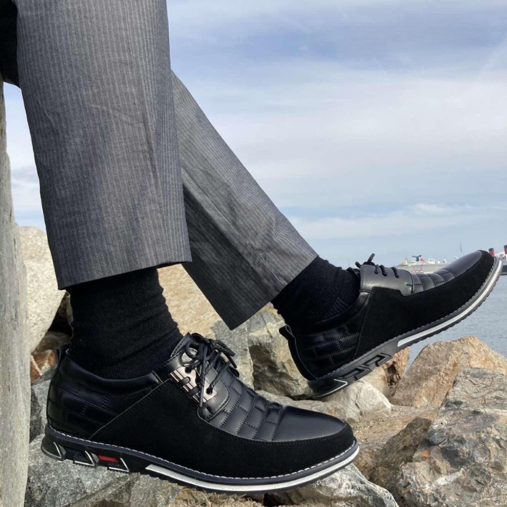 Man wearing stylish black leather sneakers with quilted design, sitting on rocks by the sea, dressed in gray pinstripe pants, showcasing casual elegance.