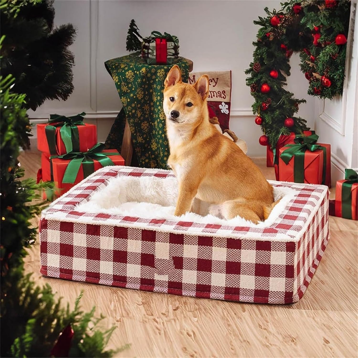 Shiba Inu dog sitting in a red plaid pet bed surrounded by Christmas decorations, gifts, and a tree, creating a festive holiday atmosphere.