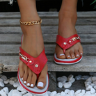 Red women's sandals with gold chain accents, worn on feet with white pedicure, resting on wooden surface surrounded by white pebbles. Fashion footwear.