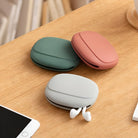 Three silicone earbud cases in green, pink, and gray on a wooden table, next to a smartphone. Ideal for earphone storage and protection.