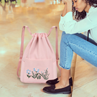 Pink drawstring backpack with floral embroidery, featuring blue and green flowers, on a marble floor next to a seated woman in casual attire.