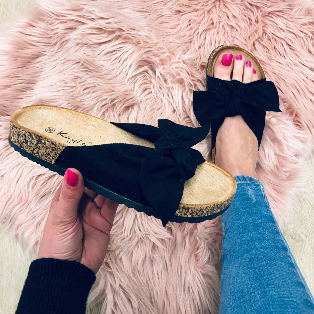 Black bow sandals with cork soles on a pink faux fur rug, worn with blue jeans. Fashionable women's footwear, casual style, summer shoes.