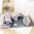 Cat playing in a pink and gray shark-shaped tunnel toy on a blue mat, with a yellow starfish toy nearby. Indoor pet activity center.