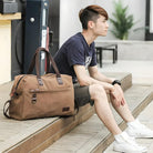 Man sitting on wooden deck with large brown leather duffel bag, wearing casual black t-shirt, shorts, and white sneakers, urban outdoor setting.