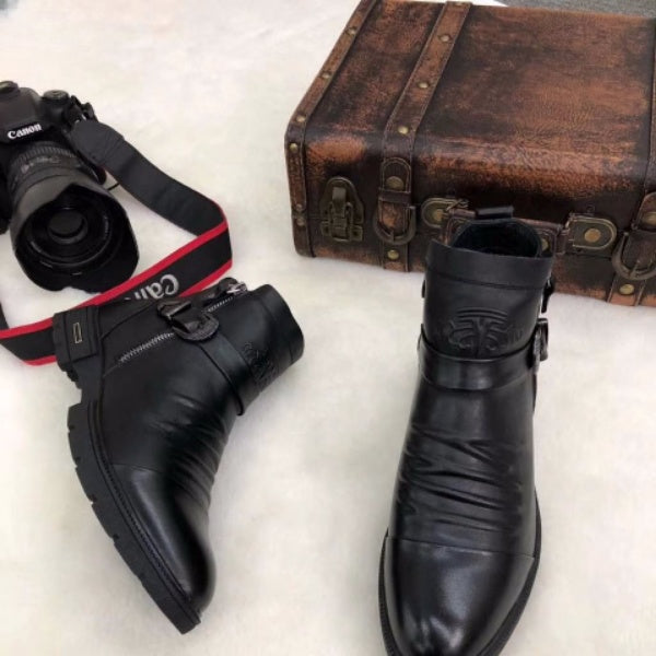 Black leather ankle boots with buckle detail next to a vintage brown suitcase and a Canon camera on a white surface. Fashion accessories display.