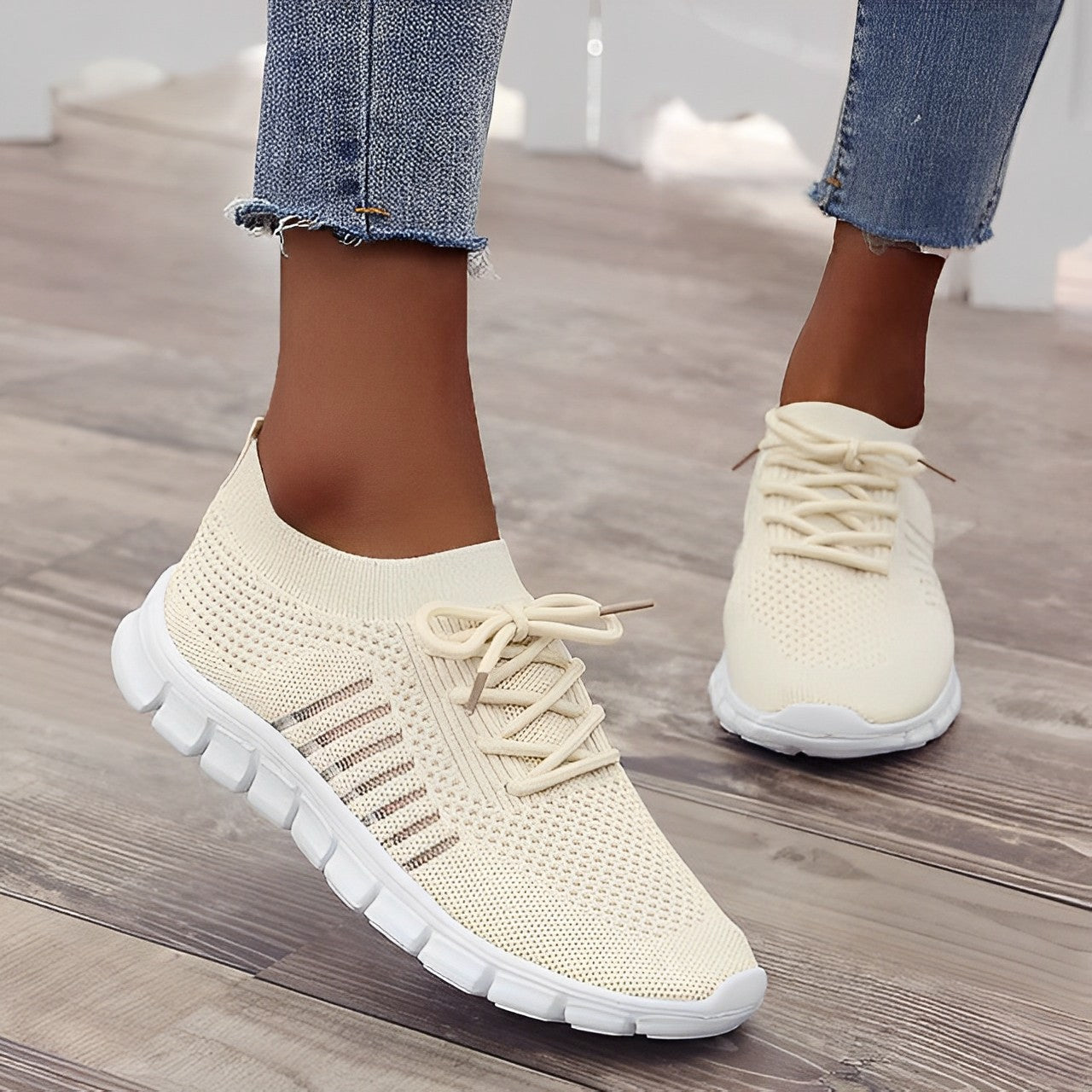 Beige knit slip-on sneakers with white soles, featuring breathable mesh design and lace-up detail, worn with distressed jeans on wooden floor.