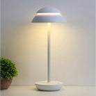 Modern white table lamp with minimalist design, featuring a dome-shaped shade and soft ambient lighting, placed on a sleek surface beside a green plant.