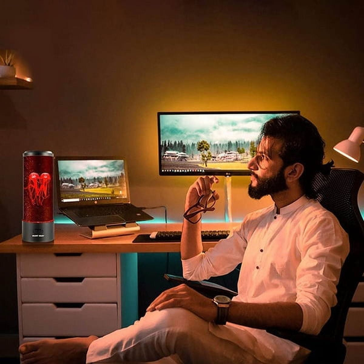 Man in a white shirt sitting at a desk with dual monitors, holding glasses, next to a red lava lamp. Cozy home office setup, warm lighting.