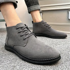 Gray suede men's ankle boots with black soles, lace-up design, worn with rolled-up jeans on a carpeted floor. Stylish casual footwear.