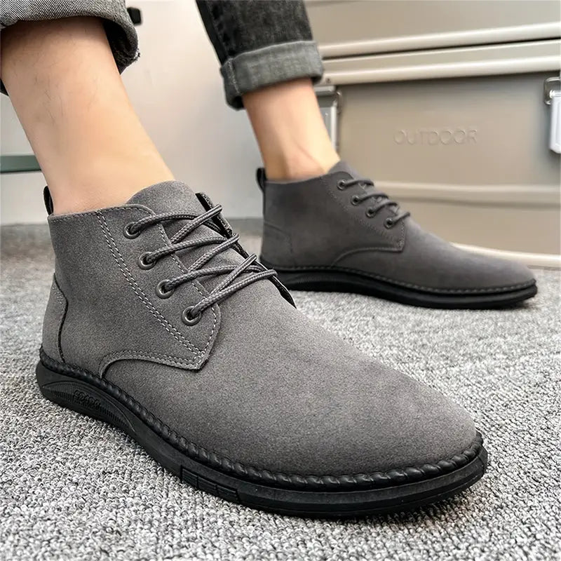 Gray suede men's ankle boots with black soles, lace-up design, worn with rolled-up jeans on a carpeted floor. Stylish casual footwear.