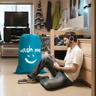 Young man in dorm room listening to music with headphones, sitting beside a large blue laundry bag labeled "wash me." Cozy student living space.