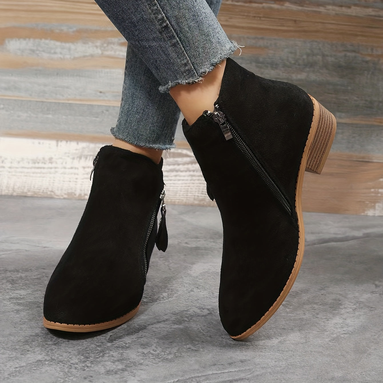 Black suede ankle boots with side zippers, worn with frayed hem jeans. Stylish women's footwear, perfect for casual and trendy fall fashion.