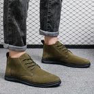 Olive green suede men's ankle boots with black soles, worn with rolled-up black jeans, on a gray carpeted floor against a wire mesh background.