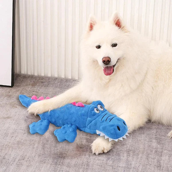 Fluffy white dog with a blue crocodile plush toy, smiling on a gray carpet. Pet toy, dog playtime, cute dog accessories, indoor pet fun.