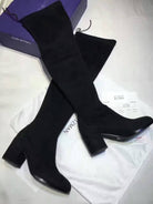 Black suede over-the-knee boots with block heels on a white background, next to a Stuart Weitzman box and dust bag. Fashion footwear.