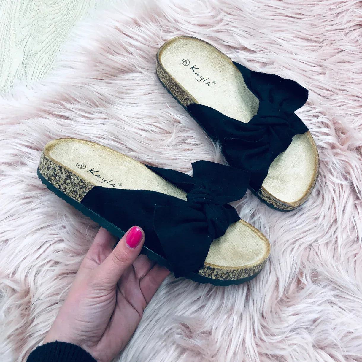 Black bow sandals with cork soles on a pink faux fur rug, held by a hand with pink nail polish. Stylish women's footwear, casual summer fashion.