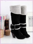 Black suede knee-high boots with gray faux fur cuffs, chunky heels, and decorative buckle, displayed on a shelf with books and wicker baskets.