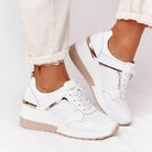 White orthopedic sneakers with gold accents, featuring a wedge heel and perforated design, worn with rolled-up beige pants. Comfortable footwear.